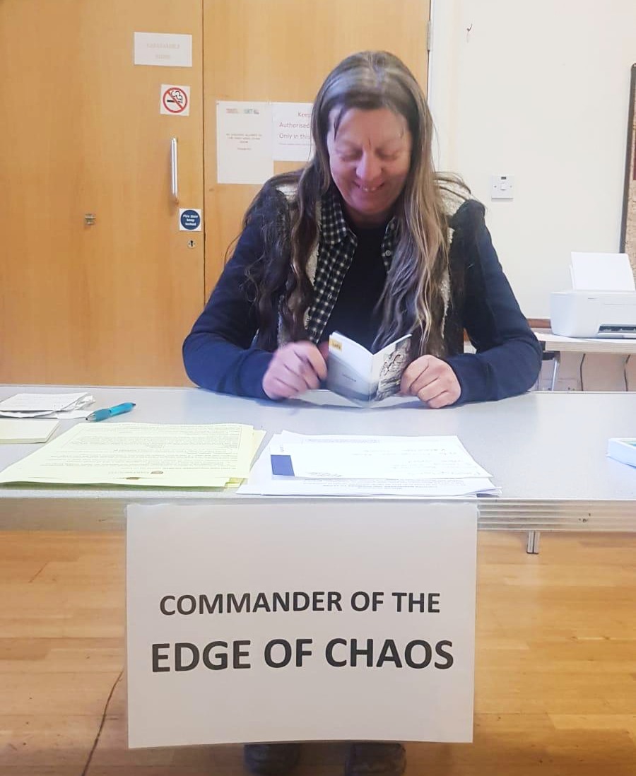 a woman is at a table reaching a notebook. Stuck to the front of the table is a piece of paper with the words 'commander of the edge of chaos'