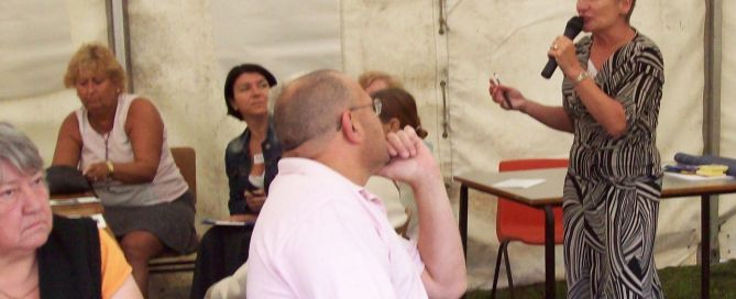 a woman is standing up holding a microphone in a marquee a number of people are sat down - some seem to be listening